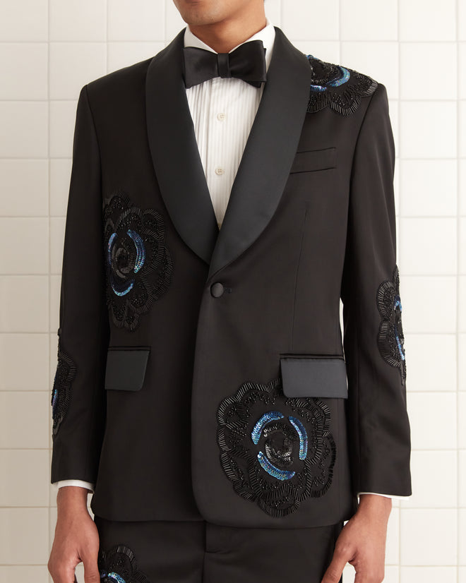 Man in a 1940s-inspired Poppy Shawl Collar black tuxedo with sequin floral patterns and beadwork, white shirt, and black bow tie, standing against a tiled wall.