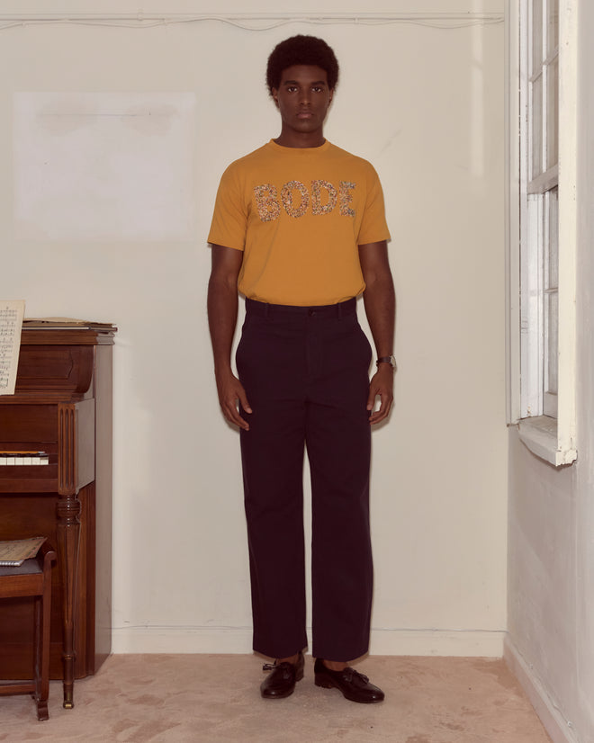 A man in a Multi Bead Tee - Yellow stands indoors next to a piano and a window, wearing black pants.