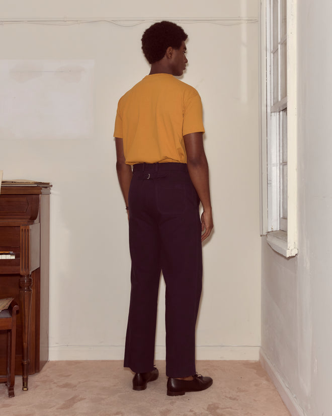 A person in a yellow Multi Bead Tee and dark pants stands facing a wall by a window, with a piano to the left.