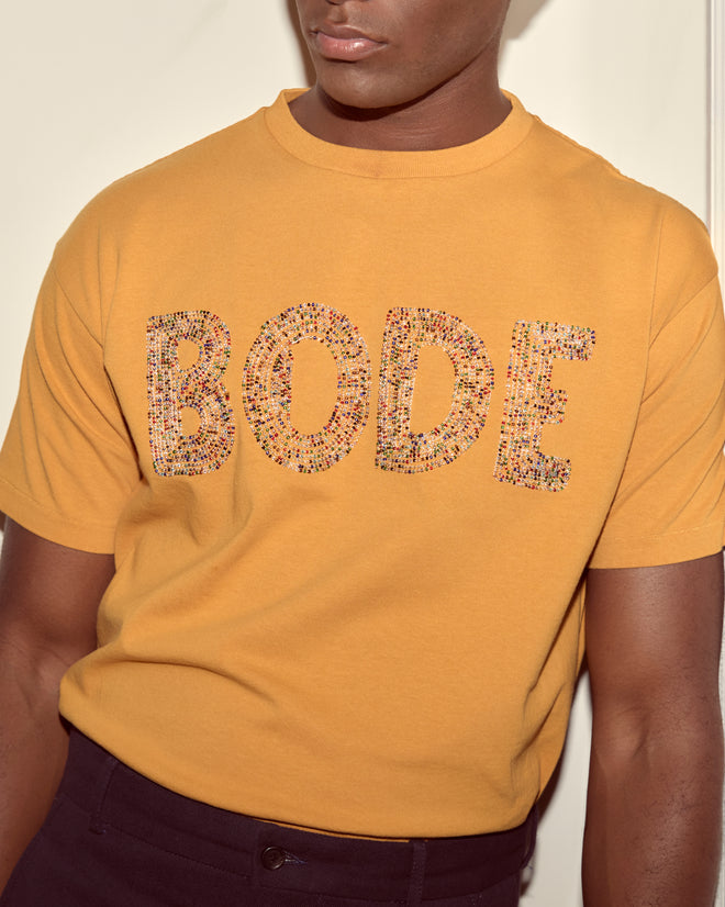 Person wearing a yellow Multi Bead Tee, made from 100% cotton and featuring “BODE” in patterned letters on the front, resembling a stylish beaded Bode T-shirt.