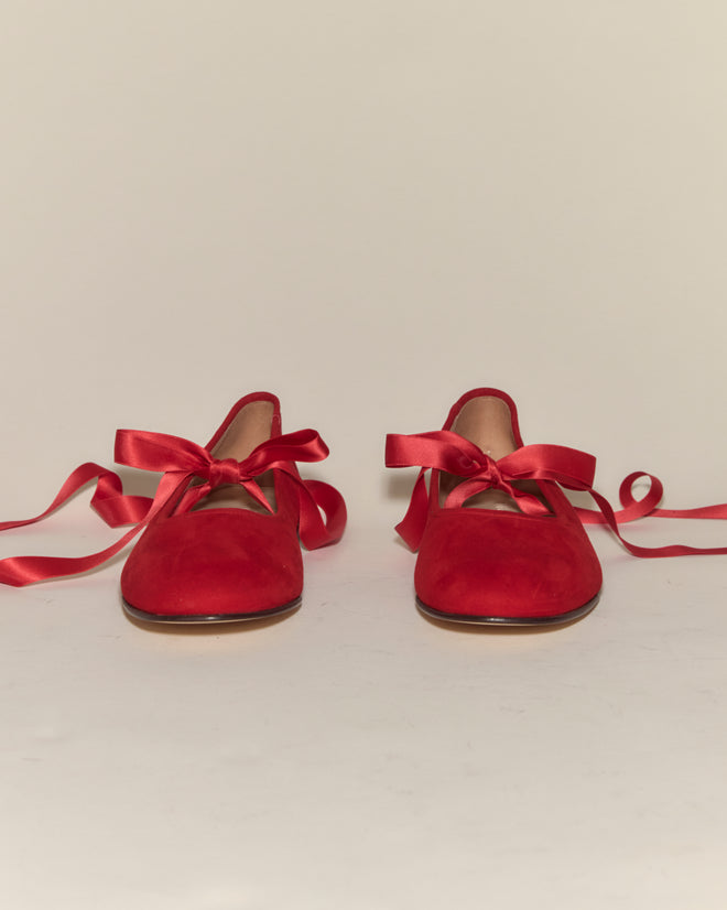 A pair of Musette Flats - Chili, red suede ballet shoes with red satin ribbon laces, placed side by side on a white surface.
