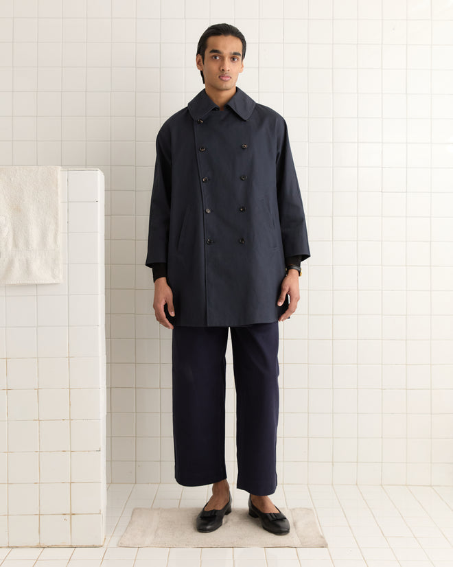 A person in a dark Waxed Herringbone Peacoat and pants stands barefoot on a rug in a tiled room.