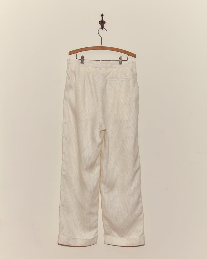 The Nelson Trousers - Ecru, a pair of light linen pants, hang on a wooden hanger against a plain wall.