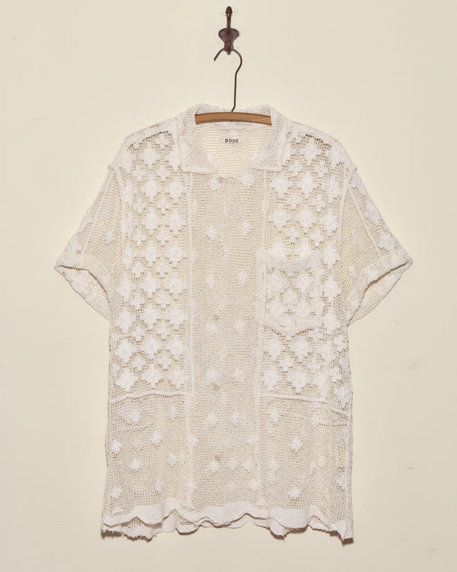 The Netted Flower Shirt - S, a unique short-sleeve white crochet lace top, is displayed on a wooden hanger against a plain wall.