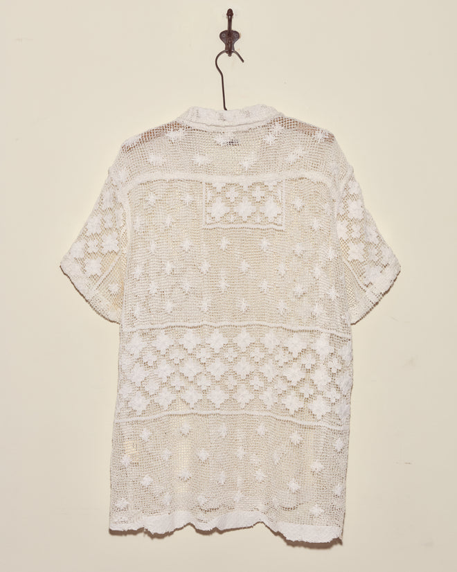 The Netted Flower Shirt - S, a unique white lace crochet piece from the 1930s with short sleeves, hangs on a hook against a plain cream wall.