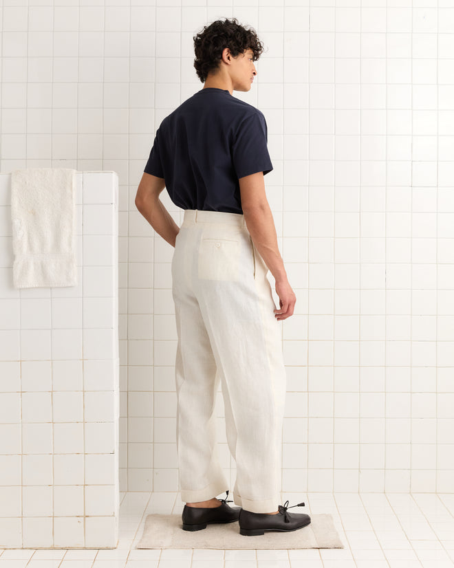 A person stands in a tiled room wearing the New England Tee - Midnight, cream pants, and black shoes, facing away.