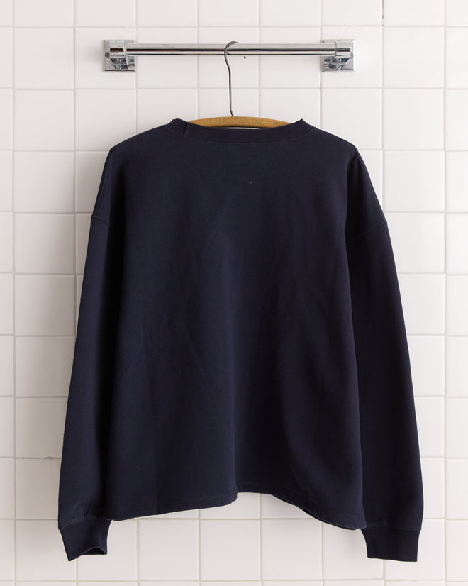 The New England Crewneck - Midnight, a 100% cotton plain black long-sleeve shirt, hangs on a wooden hanger against a white tiled wall.