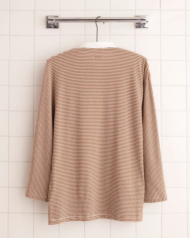 The Newton Stripe Polo - Brown, with long sleeves and a band collar, hangs elegantly on a tiled wall.