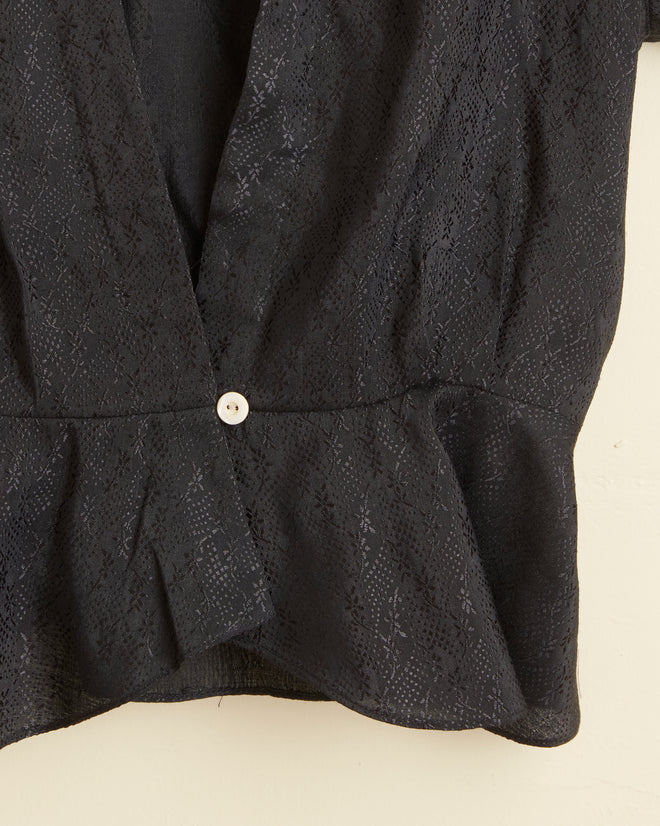 A close-up of the Night Tide Top - 10, a unique black textured blouse with a peplum hem and one white button, featuring a subtle pattern reminiscent of an early 20th-century damask tablecloth.