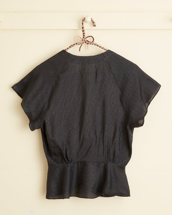The Night Tide Top - 10, a unique black blouse with short sleeves and a peplum hem, hangs on a wall hook.
