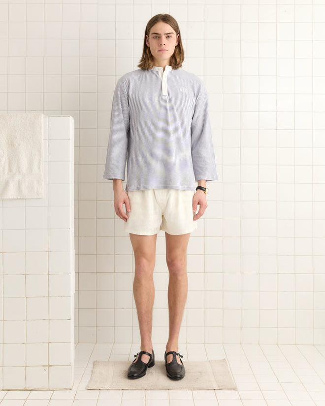 A person in a Newton Stripe Polo in light blue and white shorts stands in a tiled room with a towel nearby.