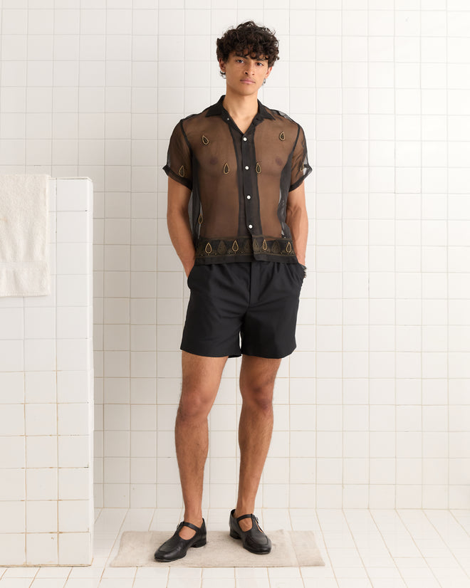 A man wearing the Pearl Paisley Short Sleeve Shirt and black shorts stands in a white tiled room.