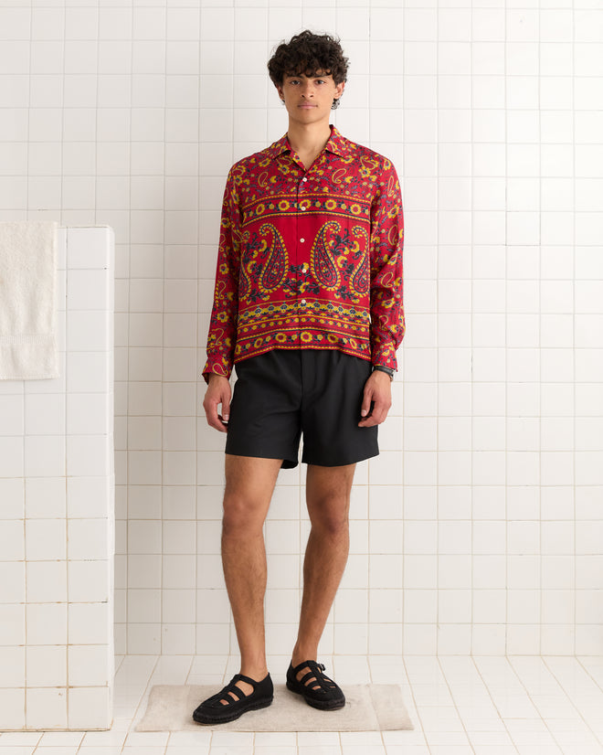 A person wearing a Cherry Paisley Long Sleeve Shirt, black shorts, and sandals stands in a white tiled room.