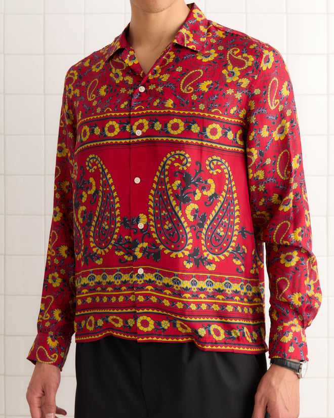 A man wears the Cherry Paisley Long Sleeve Shirt, featuring a red, yellow, and black paisley pattern made in India, as he stands against a white tiled wall.