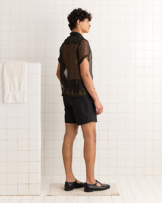 A man in the Pearl Paisley Short Sleeve Shirt and black shorts stands sideways in a tiled bathroom.