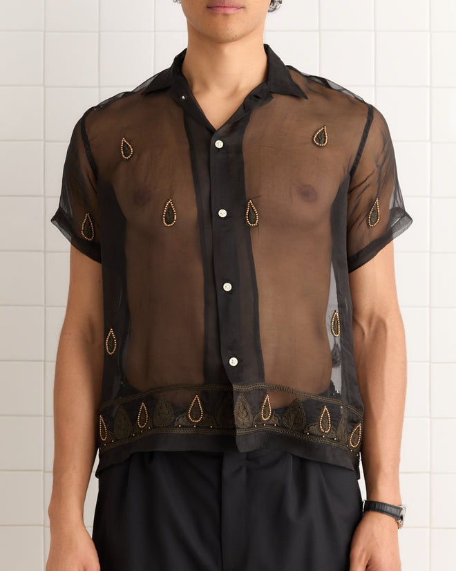 A person wearing the Pearl Paisley Short Sleeve Shirt, a sheer black top with gold teardrop embellishments and delicate satin embroidery, paired with black pants.