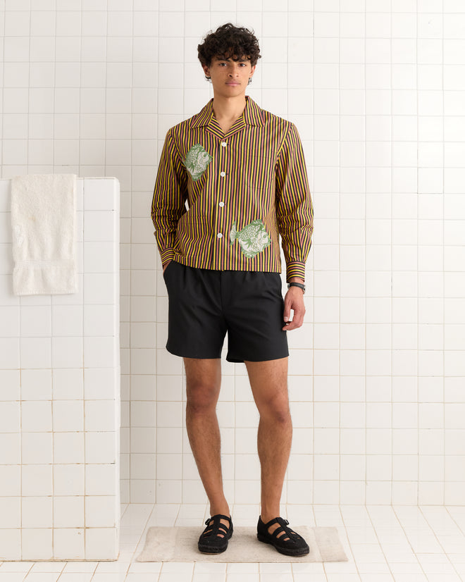 A man wearing a Fish Appliqué Long Sleeve Shirt and black shorts stands in a white tiled room beside a towel on a partition.