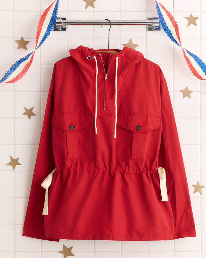 A Nylon Tie Anorak - Red hangs on a rack with festive garlands and gold stars in the background, perfect for brightening up your adventures.