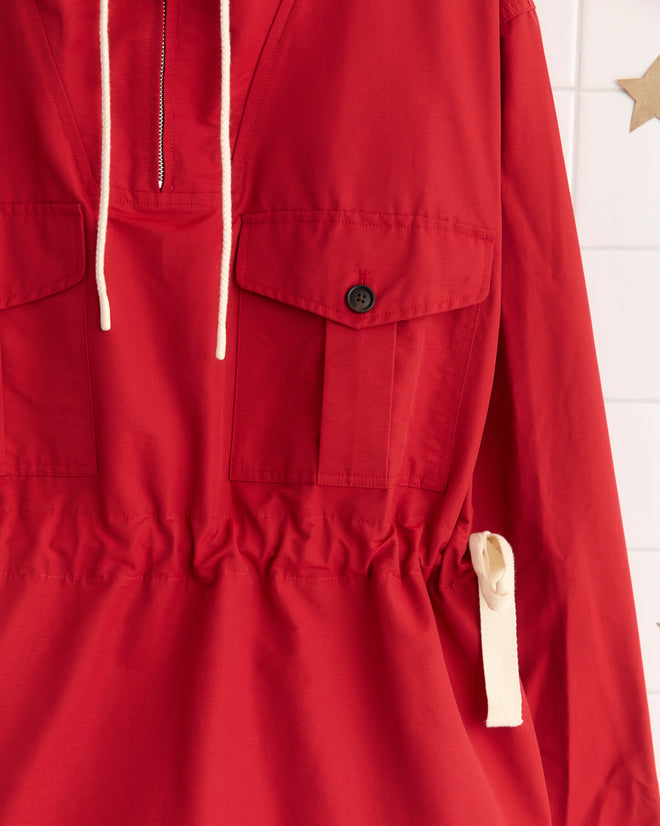 The Nylon Tie Anorak - Red features a long-sleeve pullover design with front pockets, white drawstrings, and a cinched waist with a tied belt, all crafted from durable nylon fabric.