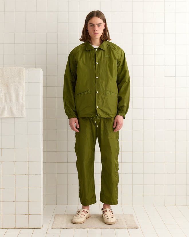 An individual wearing an Olive Nylon Track Jacket and matching pants stands in a tiled room, with a towel on the left wall.