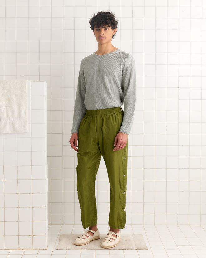 A person wearing a gray long-sleeve shirt and Nylon Tear-Away Track Pants - Olive stands in a white tiled room.