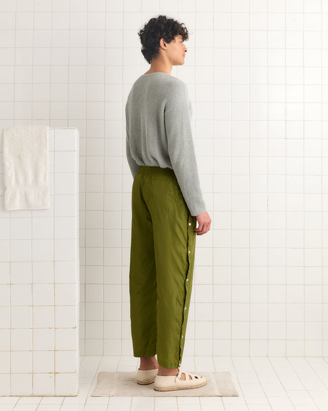 Person standing sideways in a tiled room, wearing a gray sweater, Nylon Tear-Away Track Pants - Olive, and beige shoes.