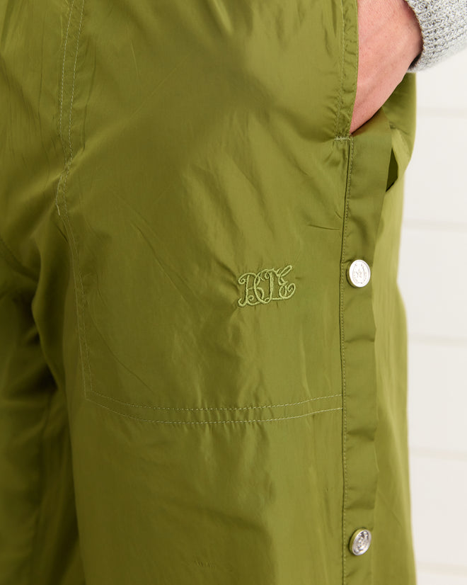 Someone wears Nylon Tear-Away Track Pants - Olive, featuring a hand in the pocket and silver side buttons, crafted from shimmery nylon.