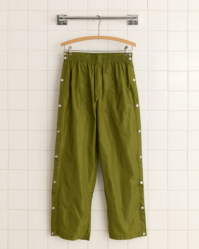 Olive Nylon Tear-Away Track Pants with side buttons hang on a hanger against a white tiled wall.