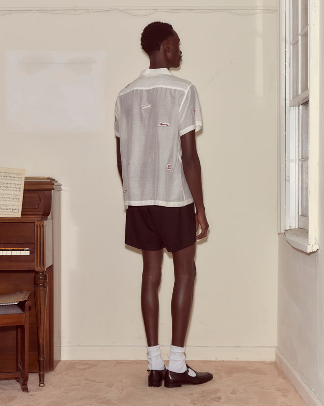 A person in the On Air Short Sleeve and black shorts stands facing a beige wall near a piano and a window.
