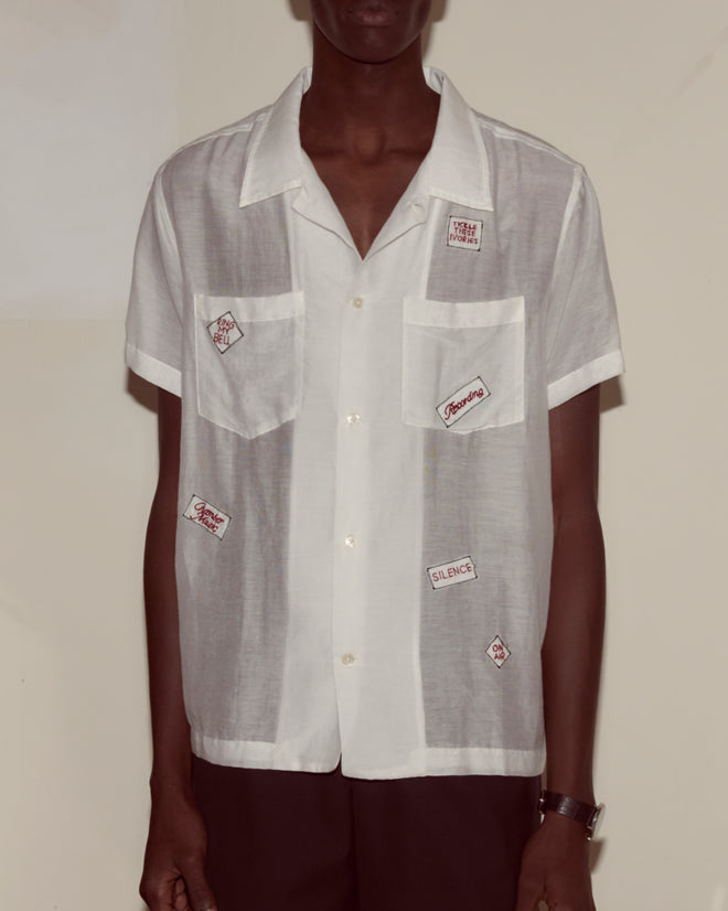 A person wears the On Air Short Sleeve, a sheer white cotton-silk shirt with embroidered patches, paired with dark pants and standing against a plain wall.