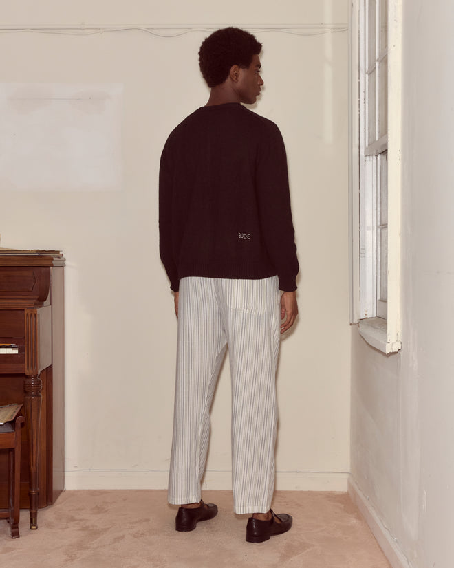 A man stands indoors by a piano and window, facing the wall while wearing the Orchard Cardigan - Black and striped pants.