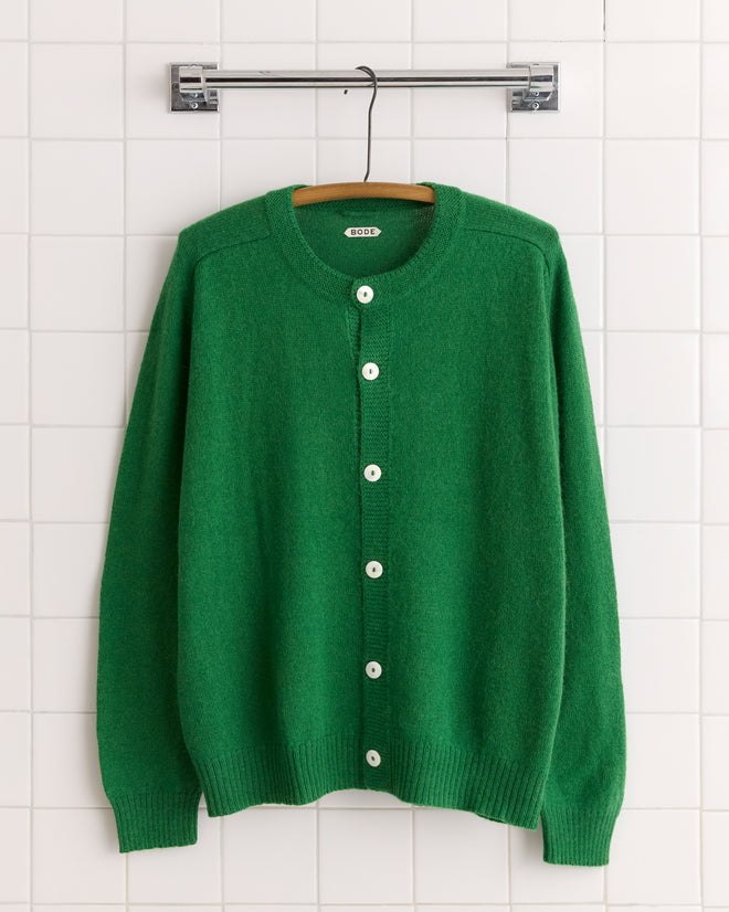 The Orchard Cardigan in Kelly Green, made from soft merino wool with antique pearl buttons, is shown hanging on a white tiled wall.