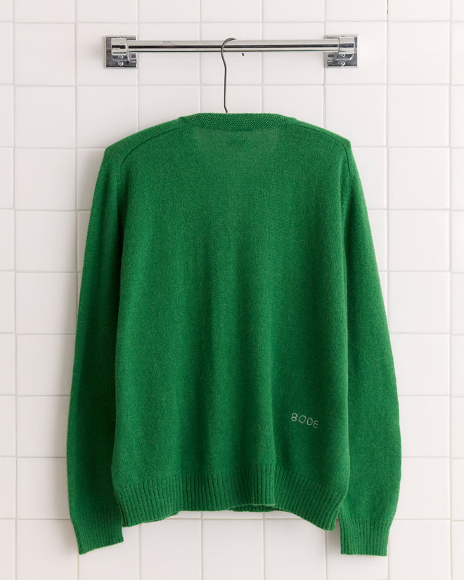 The Orchard Cardigan in Kelly Green, made from alpaca wool and featuring antique pearl buttons, hangs on a black hanger against a white tiled wall.