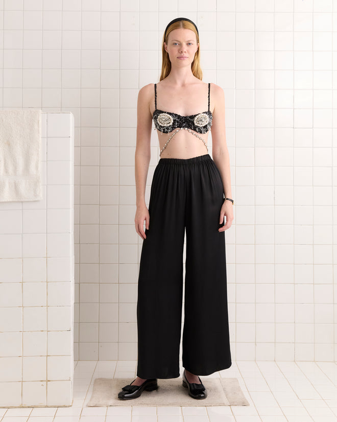 A woman stands in a tiled room wearing the Oyster Bra - Black and high-waisted black pants.