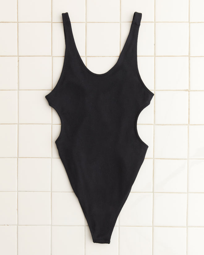 The Palmer Leotard, a sleek black one-piece with high-cut sides, is displayed flat on a white tiled surface.