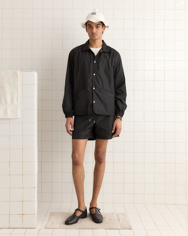 A person in a black jacket, shorts, and black shoes is standing in a tiled room wearing the elegantly embroidered Paneled Grandfather Hat in white.