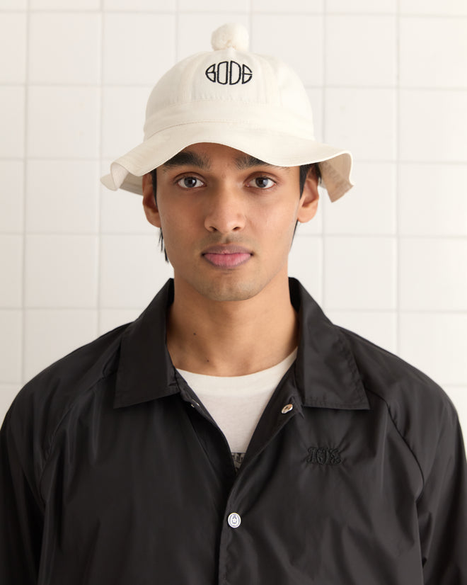 A person wearing a Paneled Grandfather Hat in white and a black jacket stands against a white tiled wall.