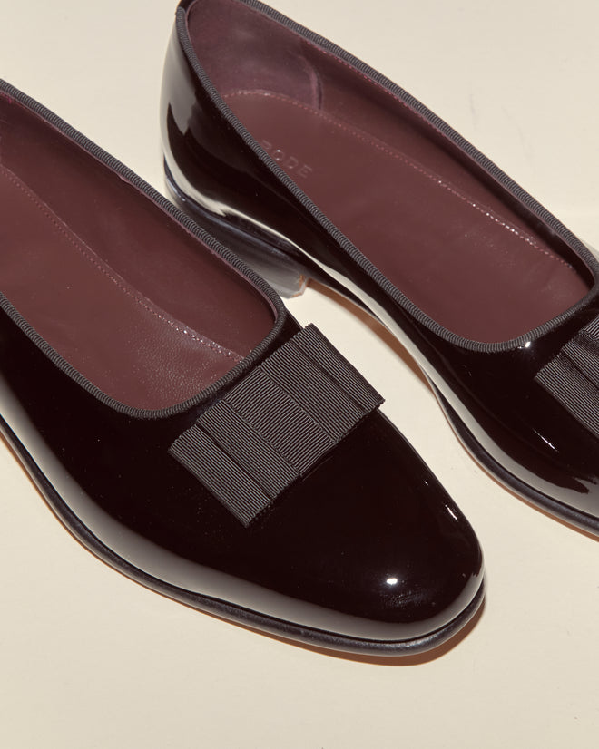 Patent Opera Pumps in shiny black patent leather with classic grosgrain ribbon bows on the toes, styled as elegant flats and photographed on a cream surface.