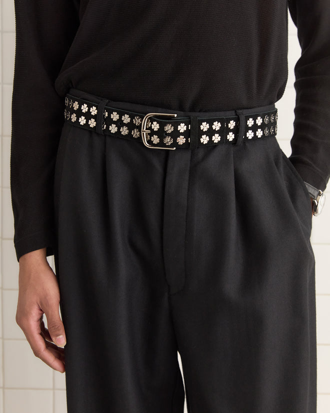 A person in a black outfit, highlighted by the Clover Charm Belt - Black, embellished with floral metal decorations, stands gracefully against a light background.
