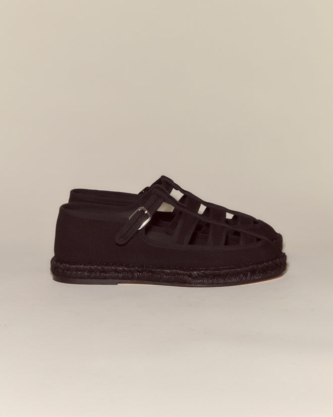 Peggy Espadrilles - Black: Black woven espadrille sandals with a jute sole and buckle, set against a plain beige background—ideal for effortless summer style.