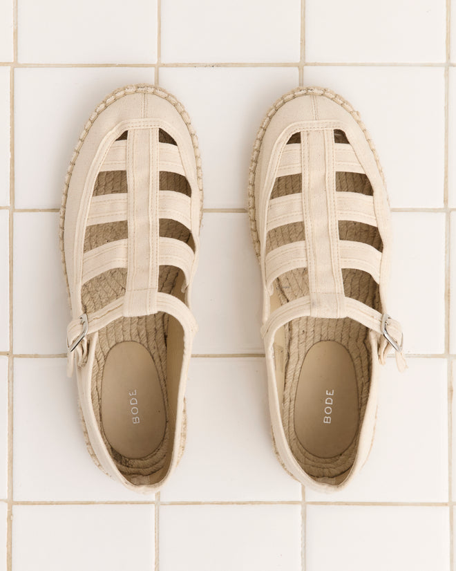 The Peggy Espadrilles - Natural feature beige woven uppers, buckle straps, and jute soles. Placed side by side on a white tiled floor, they're ideal summer shoes for effortless style.