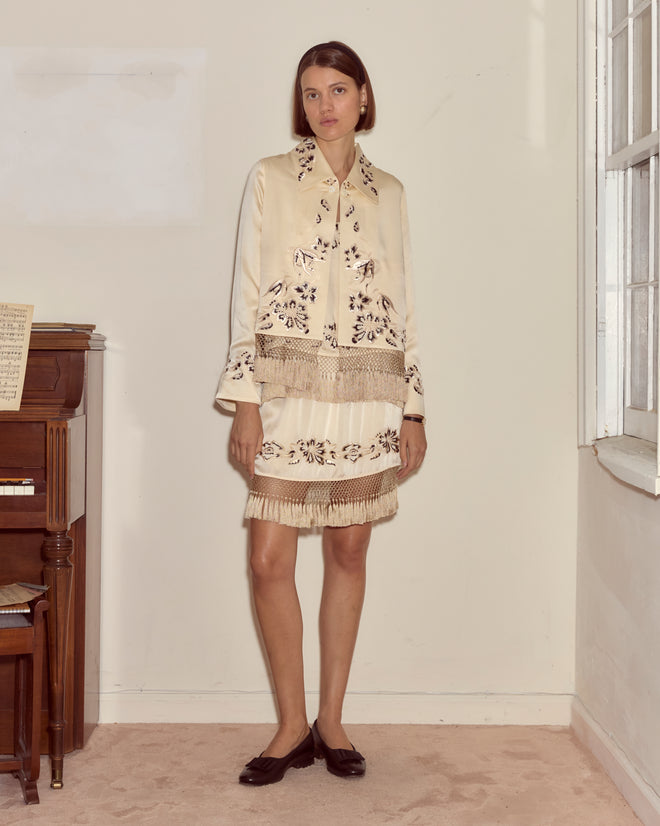 A woman in a cream floral dress and the Phantasm Fringe Jacket stands indoors by a piano and window, looking at the camera.