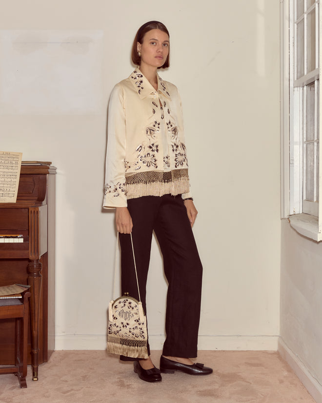 Woman stands indoors in a cream Victorian-embroidered blouse and black pants, holding the Phantasm Fringe Bag by Bode in ivory silk satin with monogram, next to a piano.