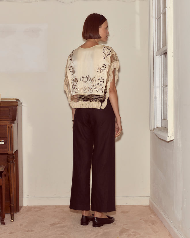 A woman with short hair stands indoors, facing away, wearing black pants and the Phantasm Fringe Shawl in cream ivory silk with delicate Victorian embroidery.