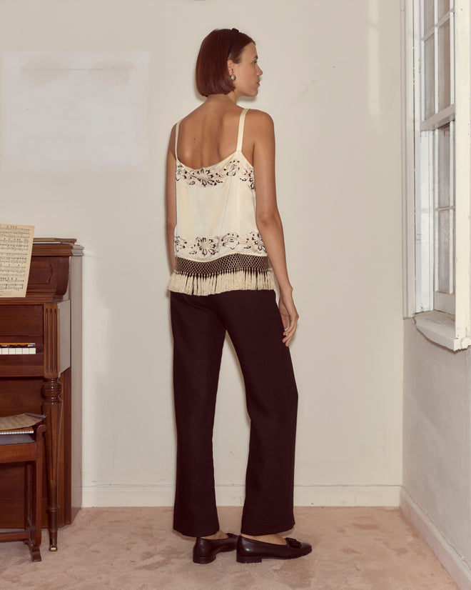 A woman in the ivory silk satin Phantasm Fringe Tank and black pants stands by a window and piano, facing away from the camera.