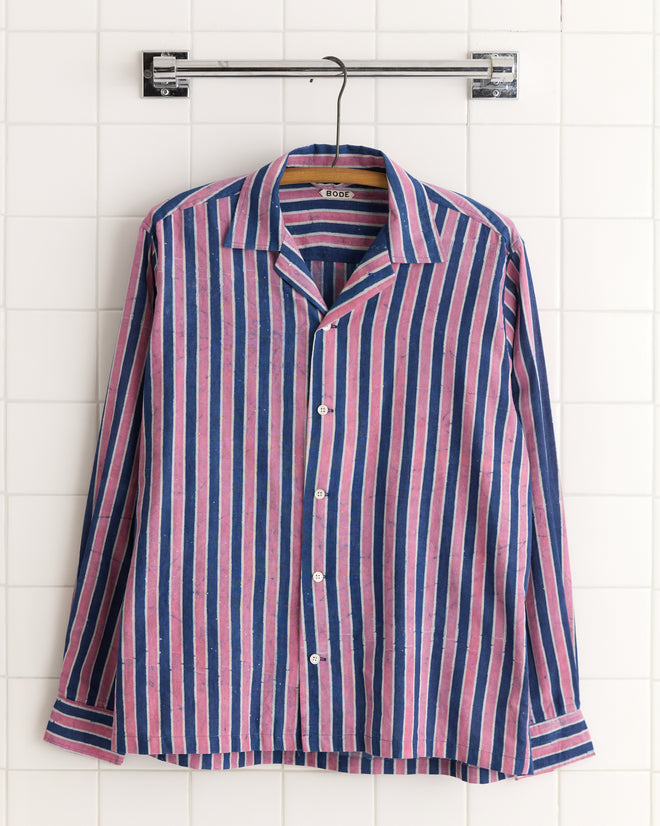 The Pink Paint Stripe Shirt, a unique long-sleeve with pink stripes, hangs on a hanger against a tiled wall.