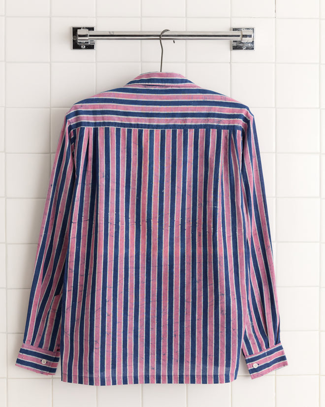 The Pink Paint Stripe Shirt, featuring unique pink, blue, and white stripes, hangs on a hanger against a white tiled wall.