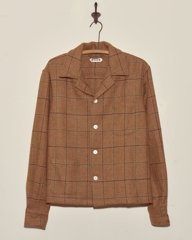The Pinscher Check Overshirt - M/L is a unique, brown plaid button-up with a collar, crafted from deadstock wool and displayed hanging on a hook against a plain, light wall.