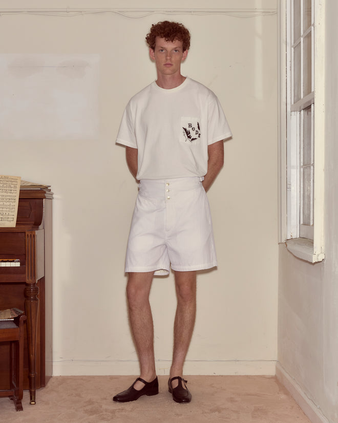 A man in a white t-shirt and Piqué Plissé Cot Shorts stands in a beige room next to a piano and a window.