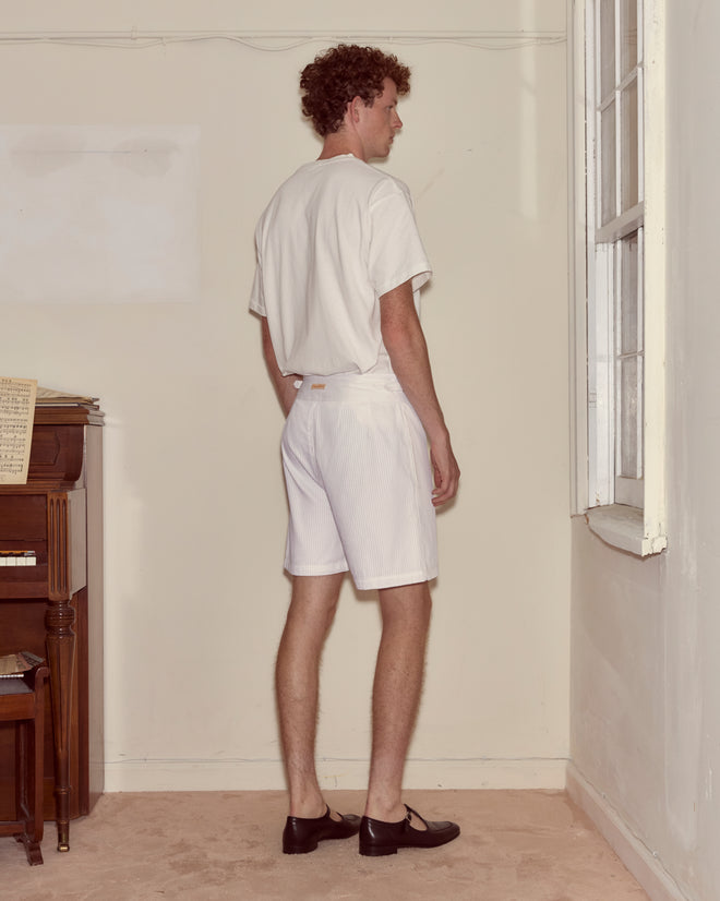 A man in a white t-shirt and Piqué Plissé Cot Shorts stands facing a corner near a window in a sparsely furnished room.